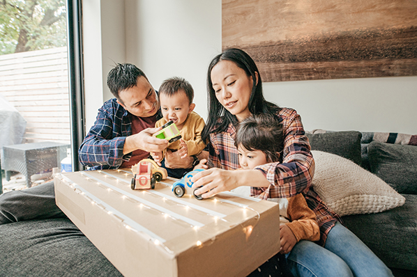 Young couple, with two young kids, playing with toy cars on a cardboard box in living room. 