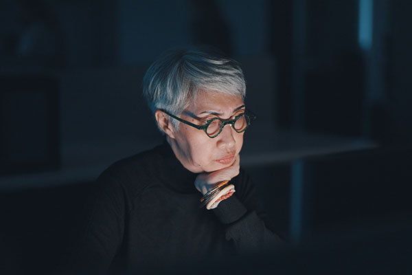 older woman of Asian descent sitting in the dark her face lit by the glow of the computer screen