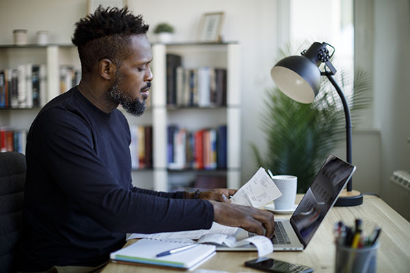 image of an adult black male using his laptop with papers in his hand and with a notebook on desk
