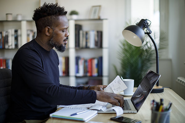 Adult male sitting at a desk reviewing his expenses while tracking them in his laptop.