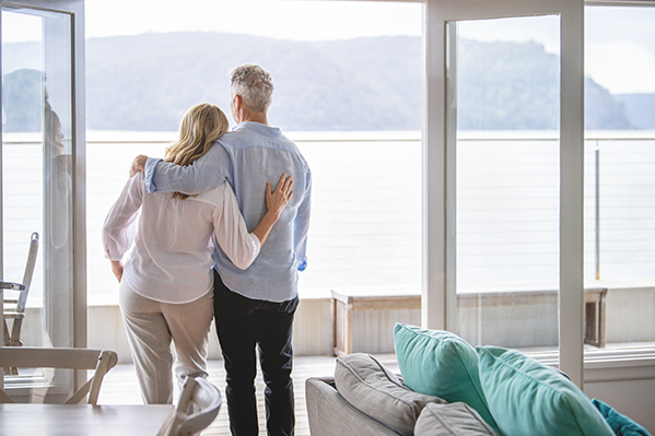 Older couple looking out the window of their retirement home with a lake and mountains in the background. 