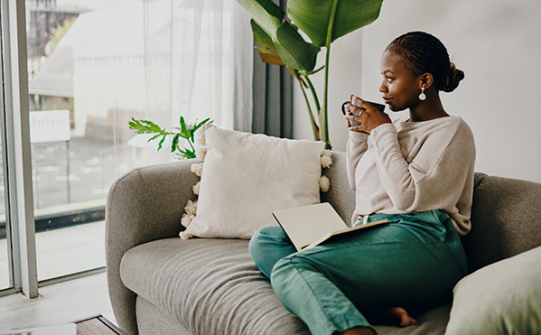 young black woman sitting on her living room couch sipping from a mug and gazing out the window