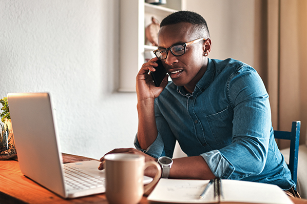 Adult male in blue shirt sitting at table. He is talking on a cell phone while looking at his laptop.